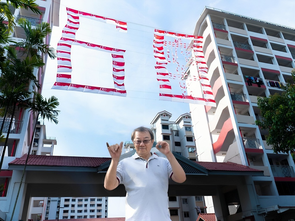 Photo of Uncle Ong standing in front of the SG60 flags, between block 107 and 109 Toa Payoh Lorong 1